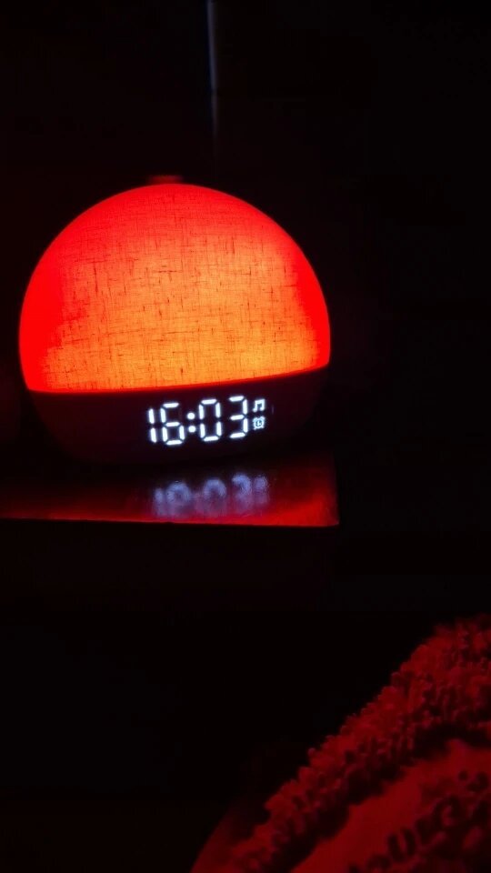 A spherical digital alarm clock glowing with a warm red-orange light, showing the time 16:03 on its front display, set against a very dark background.